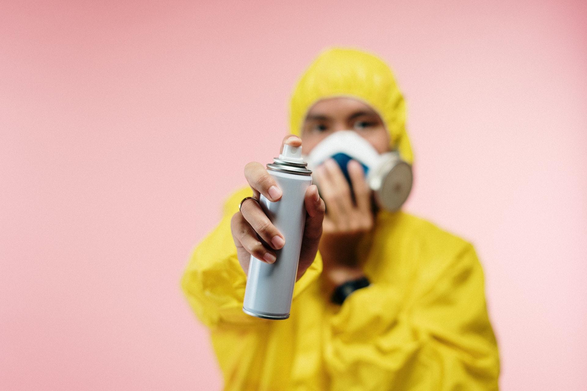 A person in a yellow hazmat suit against a peach colored wall. They are holding a respirator mask up to their face, and holding a can of aerosol spray toward the camera.