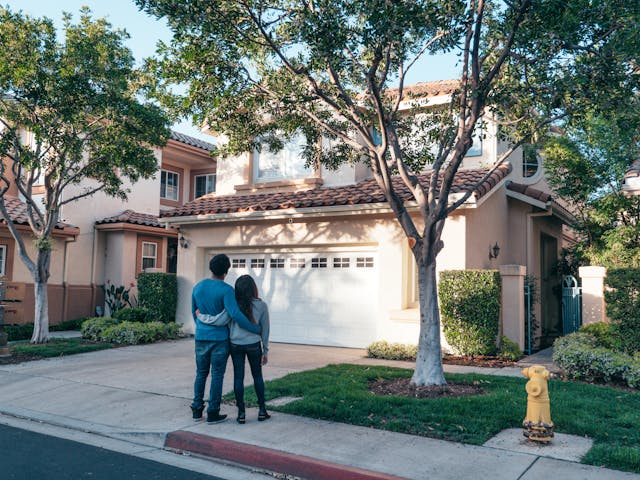 A man and woman stand with their arms around each other's waist, backs facing the camera. They are looking at a two story home from the sidewalk. The home has a young tree in it's yard, and a two car garage.