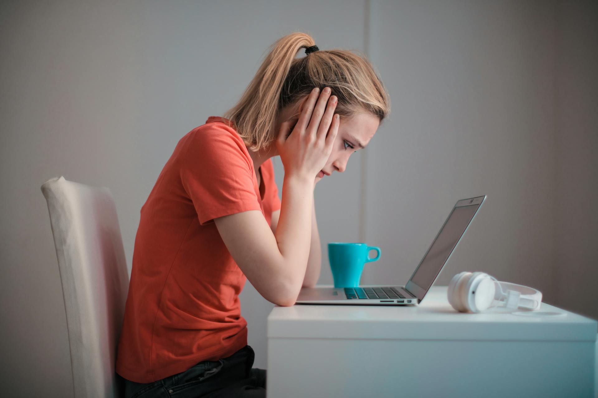 A young woman in a ponytail stares dejectedly down at her laptop with her head in her hands. She is wearing a coral t shirt. A blue mug and white headphones are also on the desk.