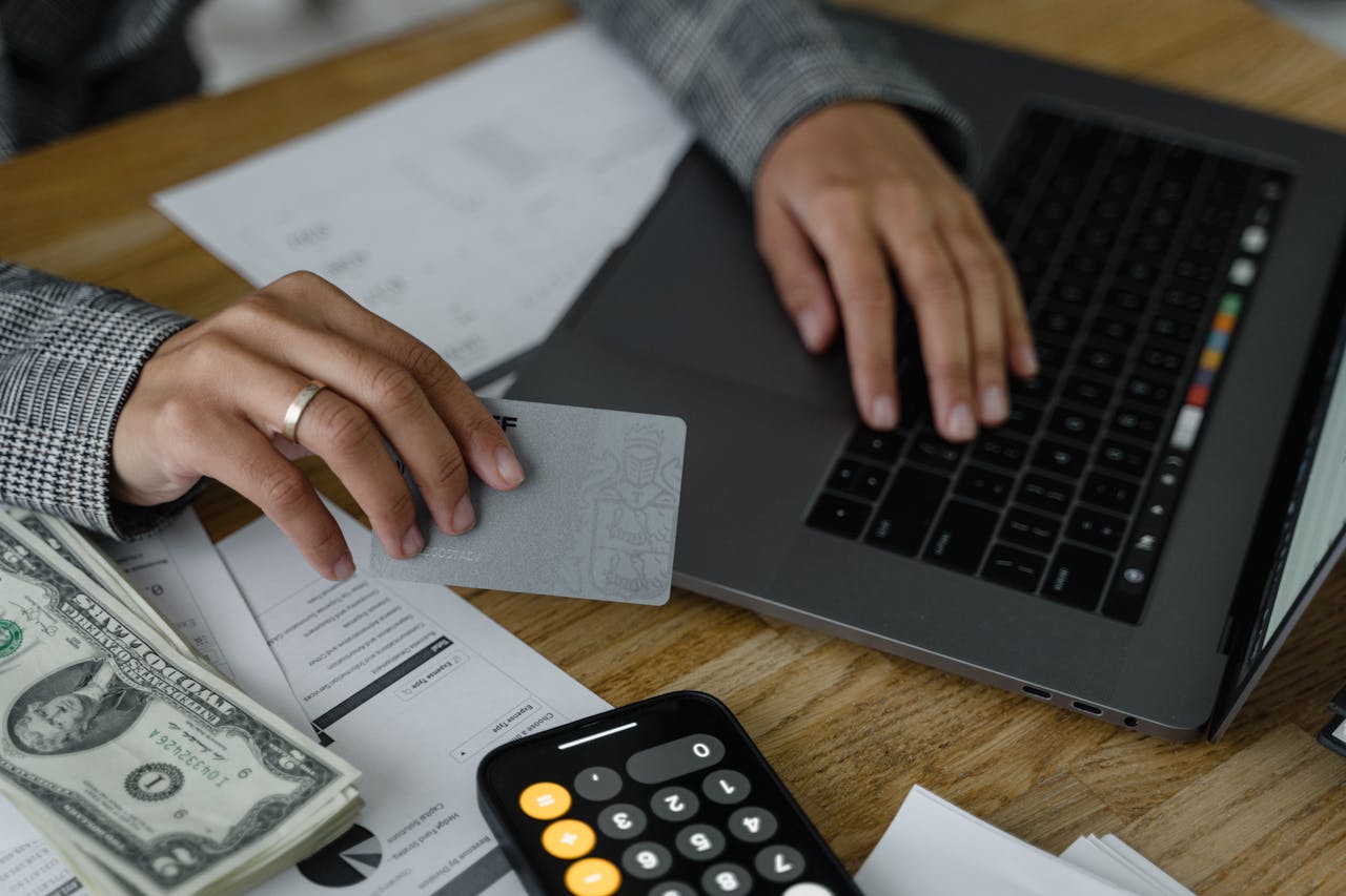 A shot of a womans hands on a desk, in one hand she holds a credit card. there is a laptop, dollar bills, documents, and a calculator