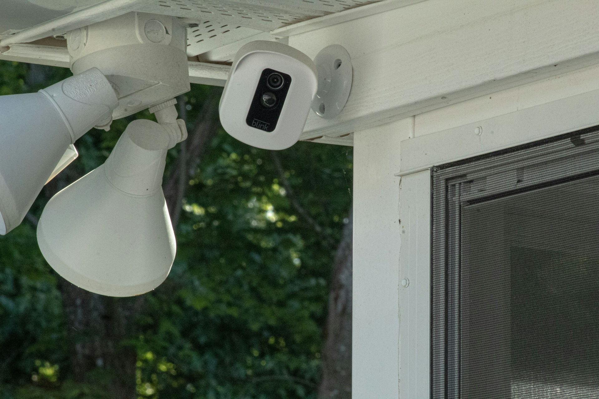 A security camera hangs on the corner of a home above a window. Behind it are two motion activated lights pointing away toward the yard.