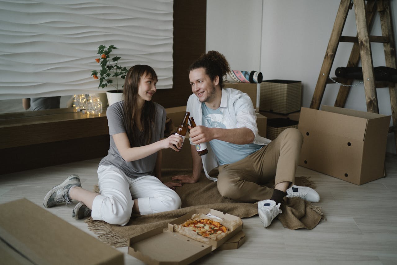 A couple sits on the floor of an apartment and toasts with two beer bottles. An open pizza box is on the floor between them.