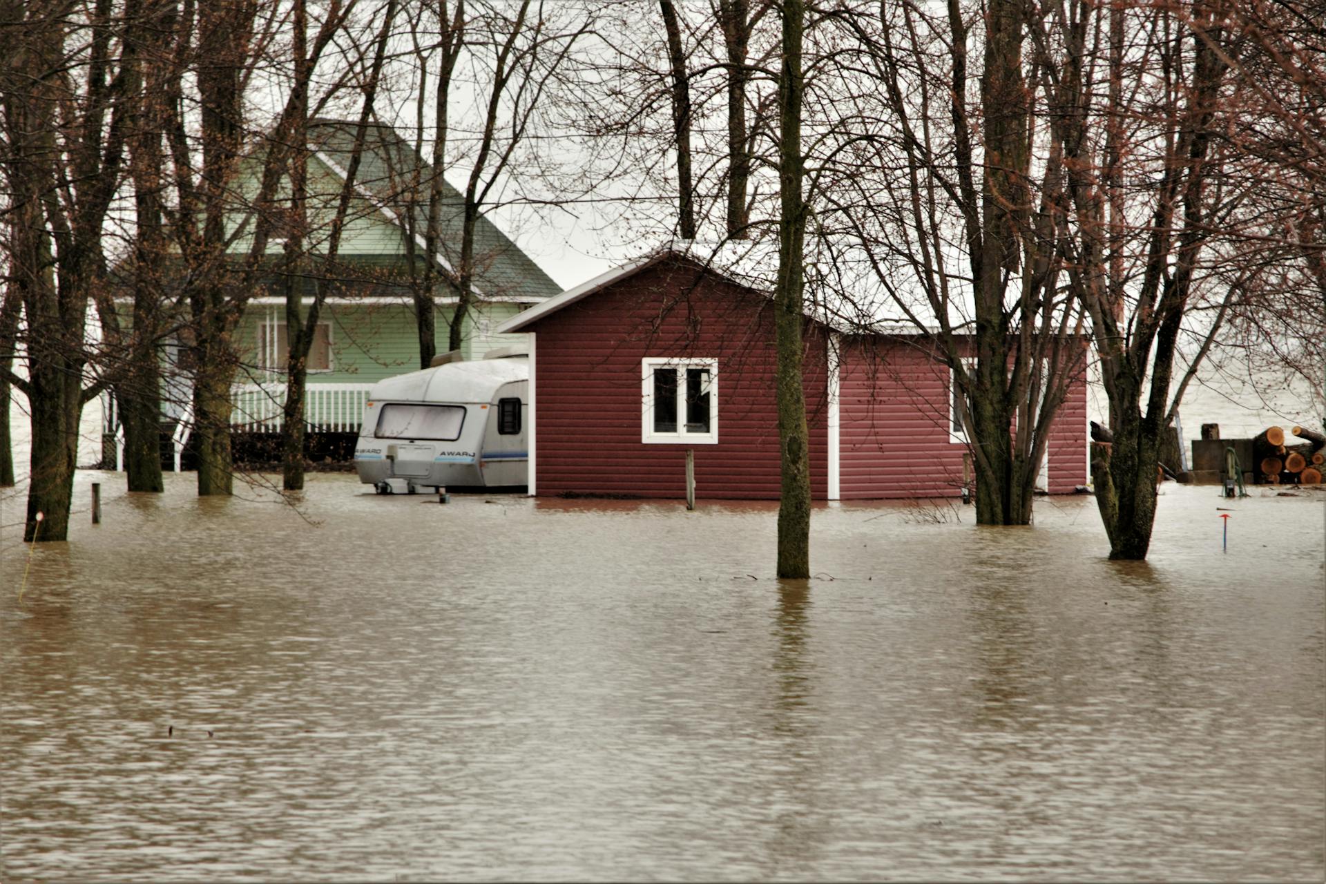 A large red shed, camping trailer, and modest home sit between leafless trees. The area is heavily flooded and water covers the ground entirely.
