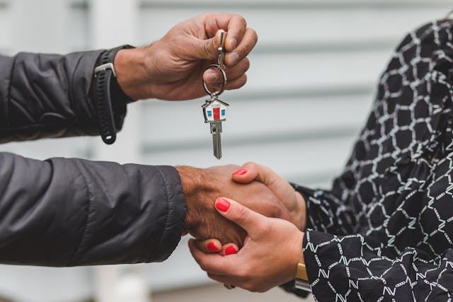 A woman with red nails grasps a man's hand in a firm handshake with both hands. The man holds out a key with the design of a home on the keychain.