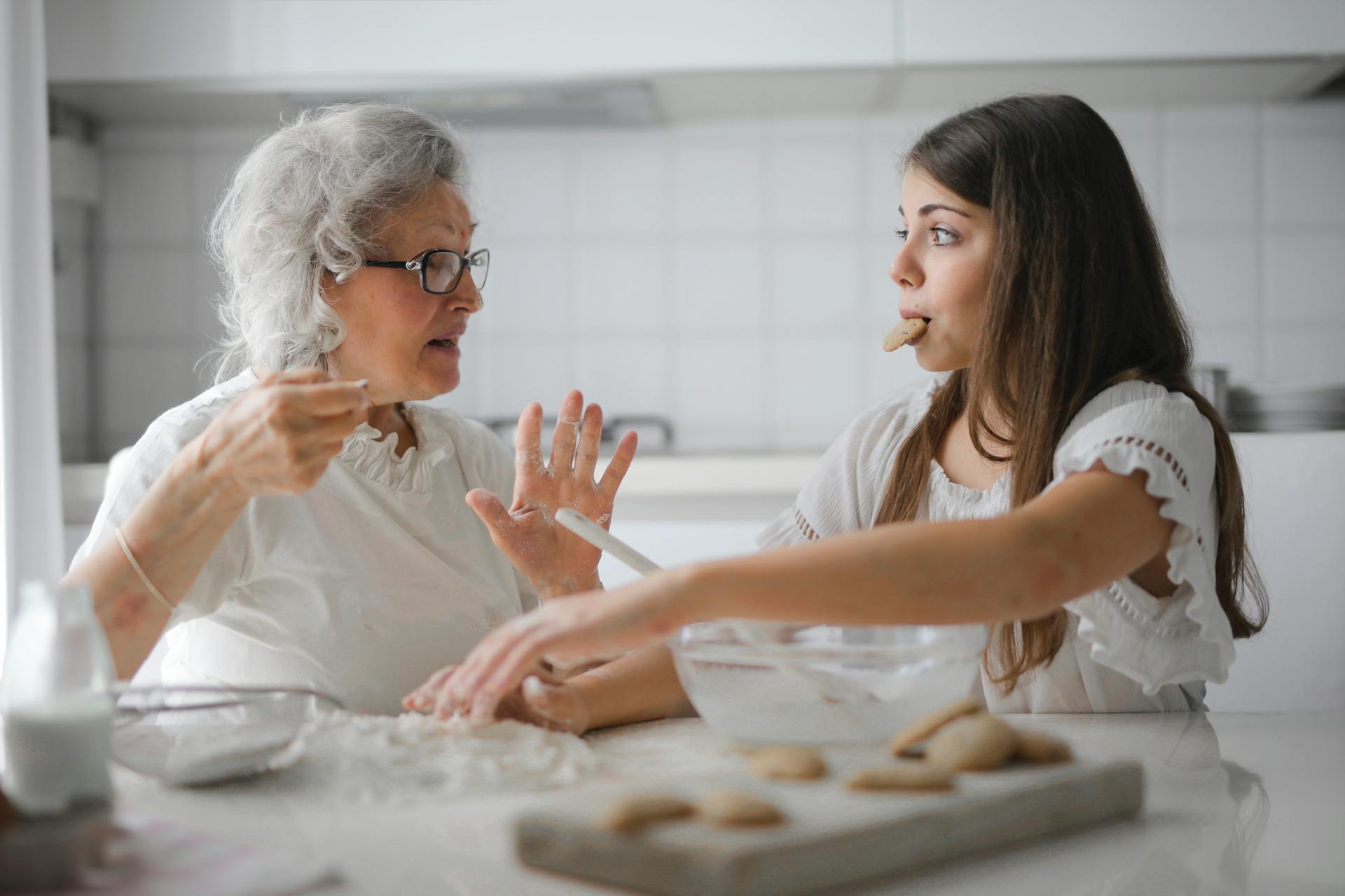 An older woman and a teenage girl sit in a clean, white kitchen. The teenager has a cookie in her mouth and is reaching for something unseen on the counter. The older woman holds her hands up as if in conversation with the younger girl.