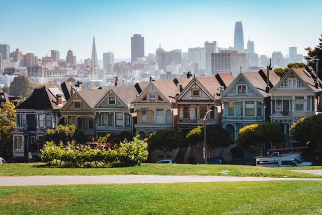 A San Francisco neighborhood. A row of duplex style homes line the street across from a vibrant green lawn. In the far distance sits the city skyline.