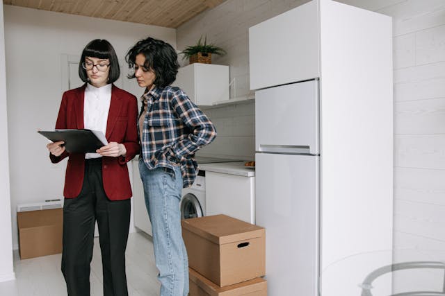 A woman in a red blazer and black slacks stands in an empty kitchen next to another woman in jeans and a flannel shirt. They are looking at paper in a portfolio in the first woman's hands. here are moving boxes on the ground.