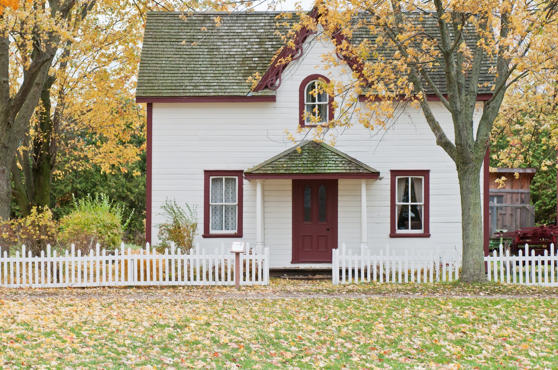 A small two story cottage style home sits between several trees. It is fall, and the leaves of the trees are yellow. The lawn has a light coating of yellow leaves.