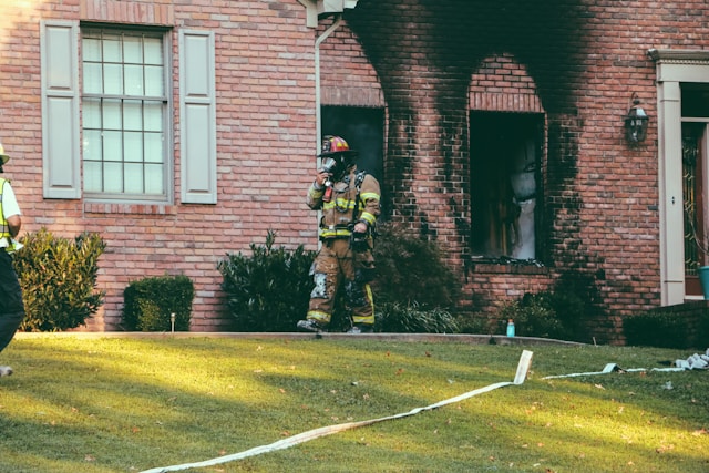 A firefighter walks along the lawn of a red brick house. Two of the windows are broken and large black scorch marks mar the bricks surrounding the broken windows.