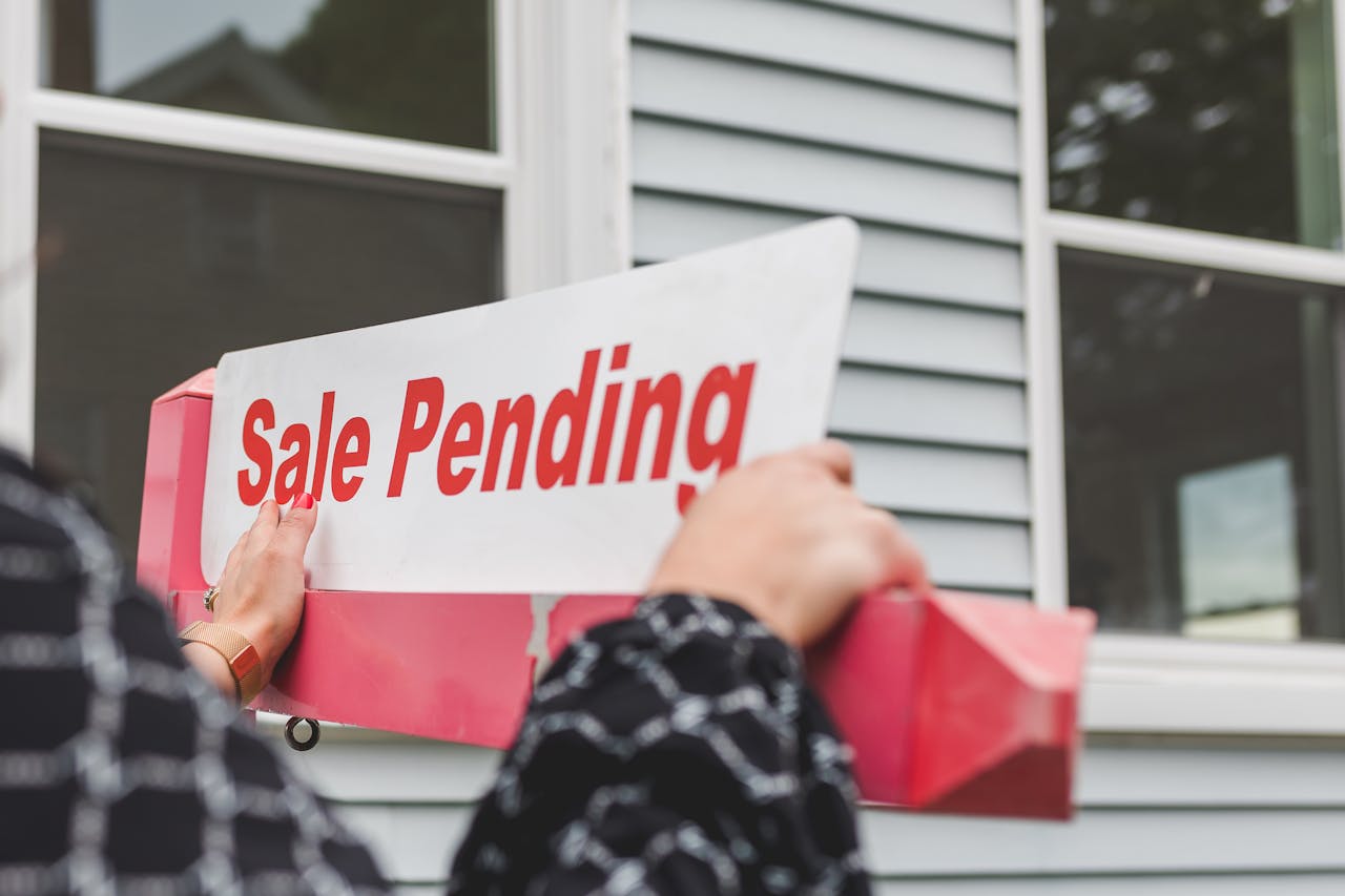 A close shot of a woman placing a Sale Pending sign in front of a home. There are two windows visible in the background.