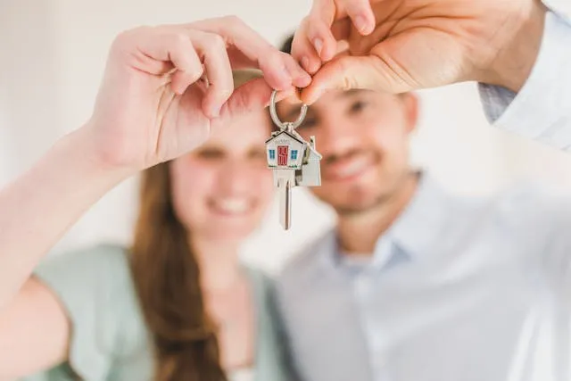 A couple holds a key with a house keychain out to the camera. Their hands and the keys are in focus, the couple is out of focus. They are smiling.