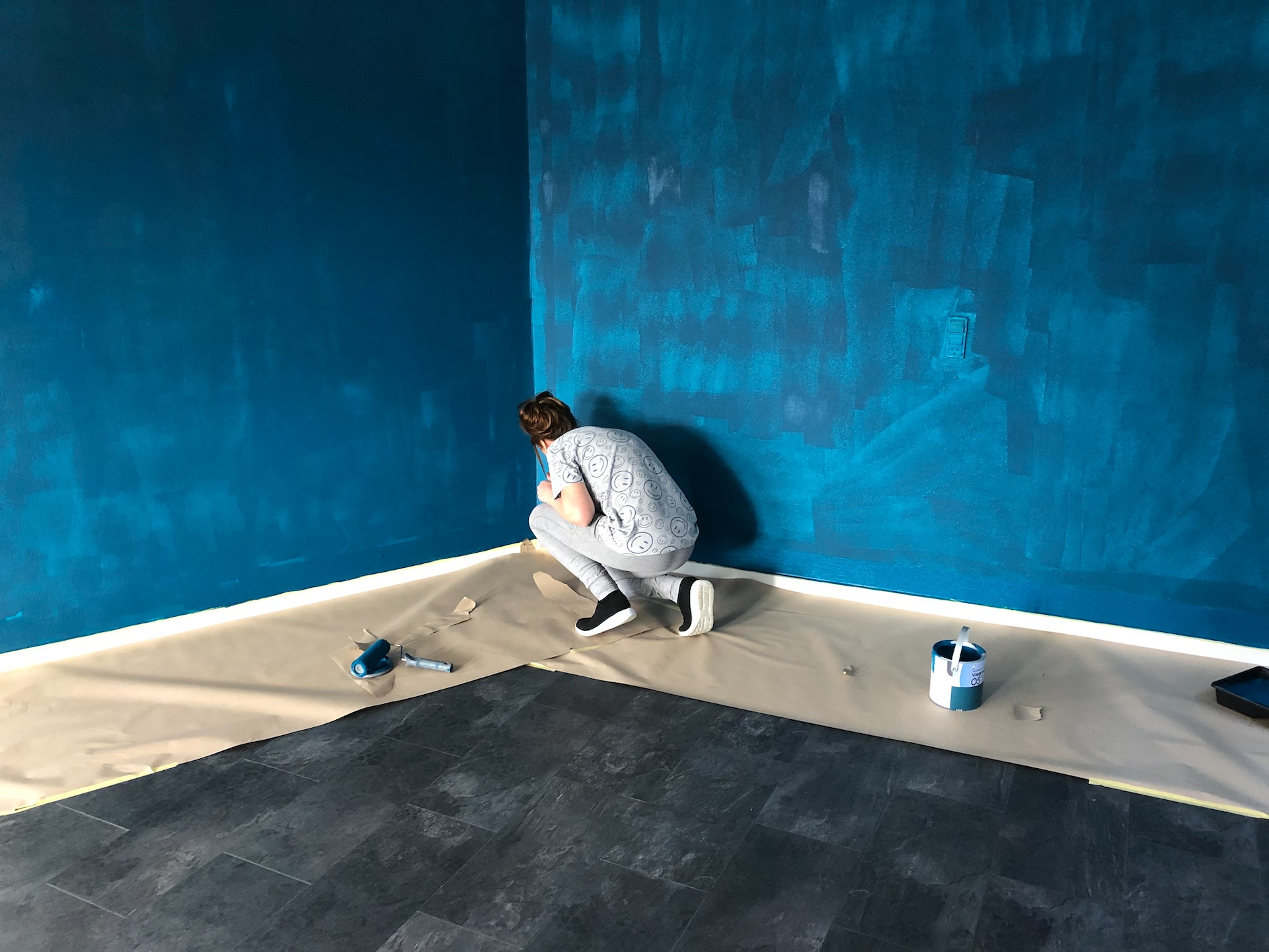 A person is crouched in a corner, detailing a paint job. The walls are a deep cerulean blue and need a few more coats. There is paper placed on the floor to protect the deep grey charcoal tile from errant paint splatter.