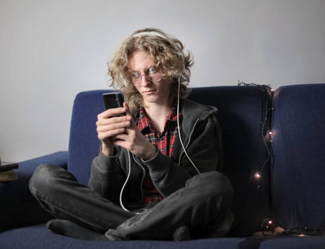 A young androgynous person sits cross-legged on a navy blue couch. They are wearing headphones and looking at a phone they hold in their hands. A string of fairy lights is draped over the back of the couch.