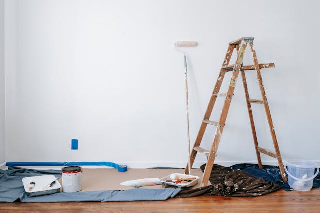 A display of various gear and tools used for interior home painting against a white wall. A ladder, paint roller, paint bucket, tape, tarp and other tools are visible.
