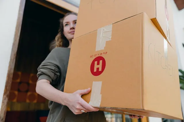 A woman wearing a gray sweatshirt with the sleeves pushed up carries two moving boxes out of a doorway. One of the boxes is labeled kitchen.