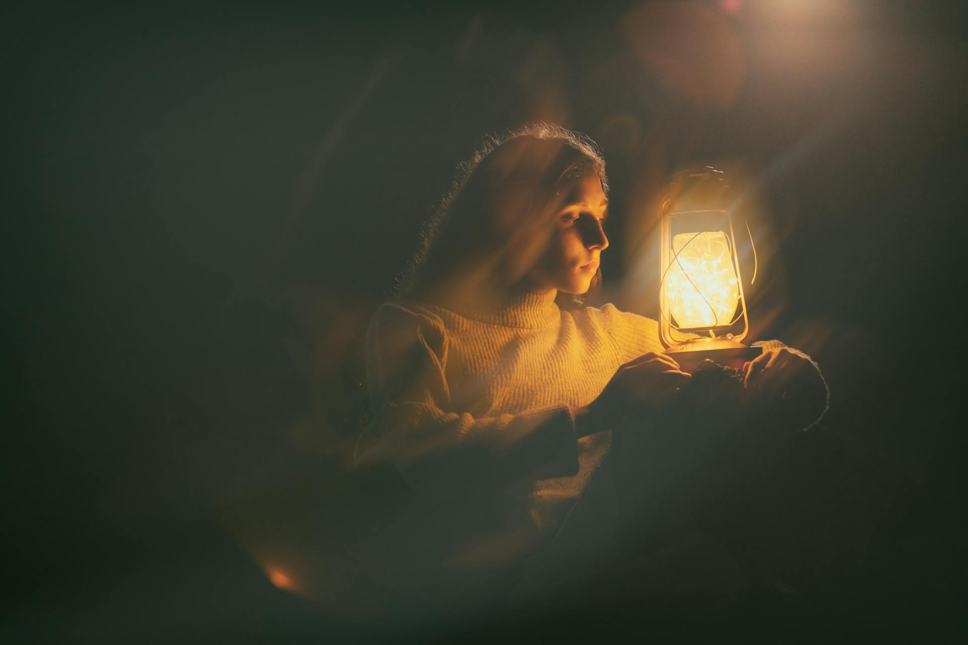 A woman sits on an unseen seat and holds a lantern. She is gazing into it with a neutral expression. Most of the photo is dark, and there are some lighting streaks and effects.