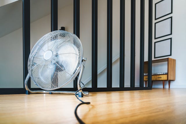 A medium sized metal fan on a wooden floor. Behind the fan are decorative black railing bars obscuring a staircase and picture frames.