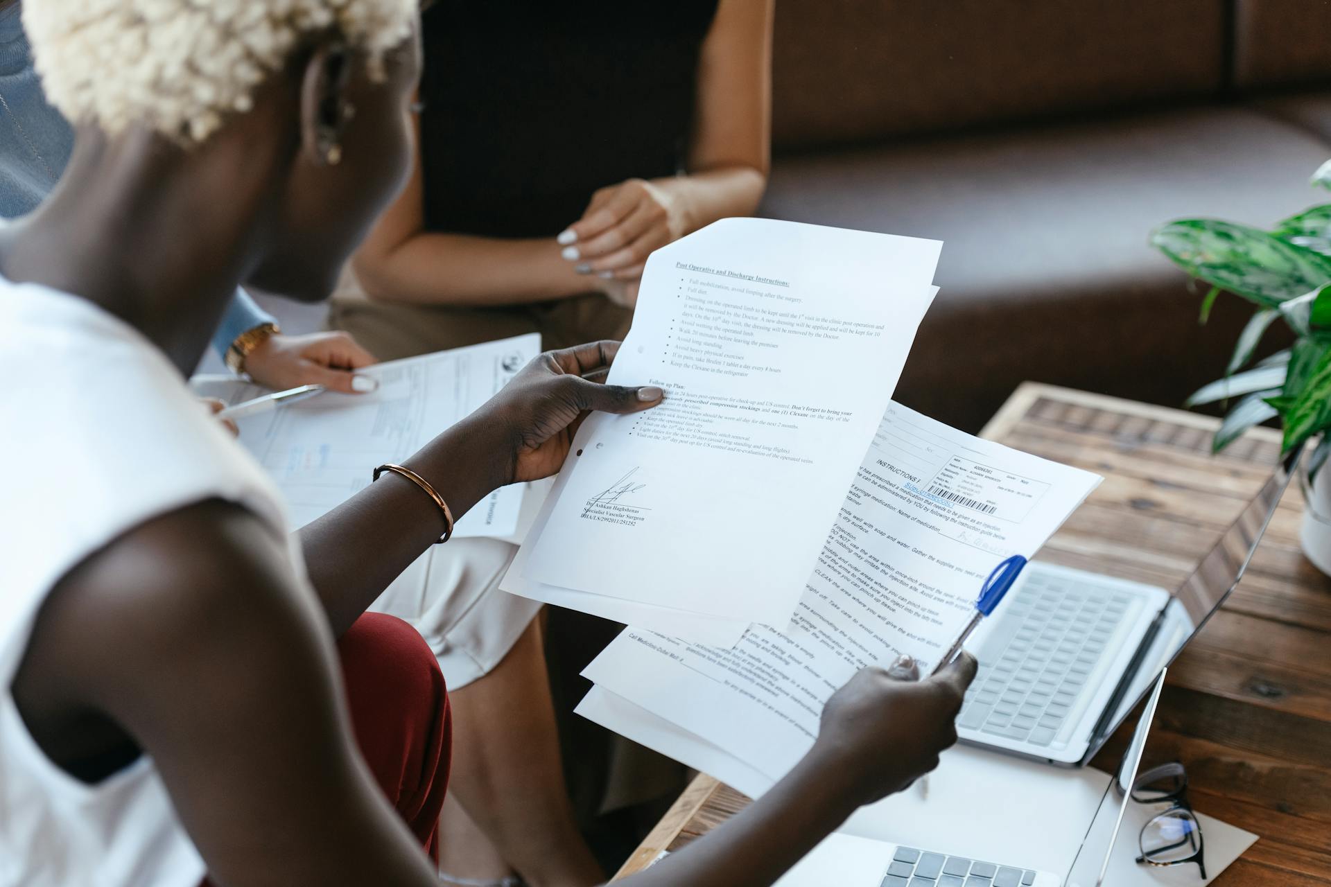A woman looks over a handful of forms and documents. The angle is over her shoulder and focused on the documents. Two other people are seated in the background, although only their arms or legs are visible.