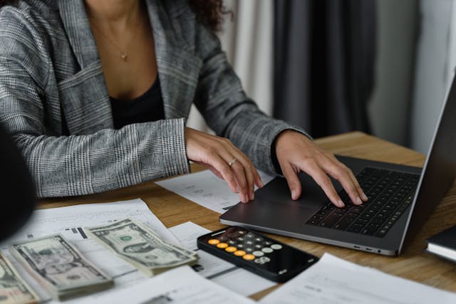 A tight framed photo of a woman working at a desk. She is wearing a gray patterned blazer and modest jewelry. On the desk are a laptop, a calculator, paperwork, and small stacks of dollar bills.