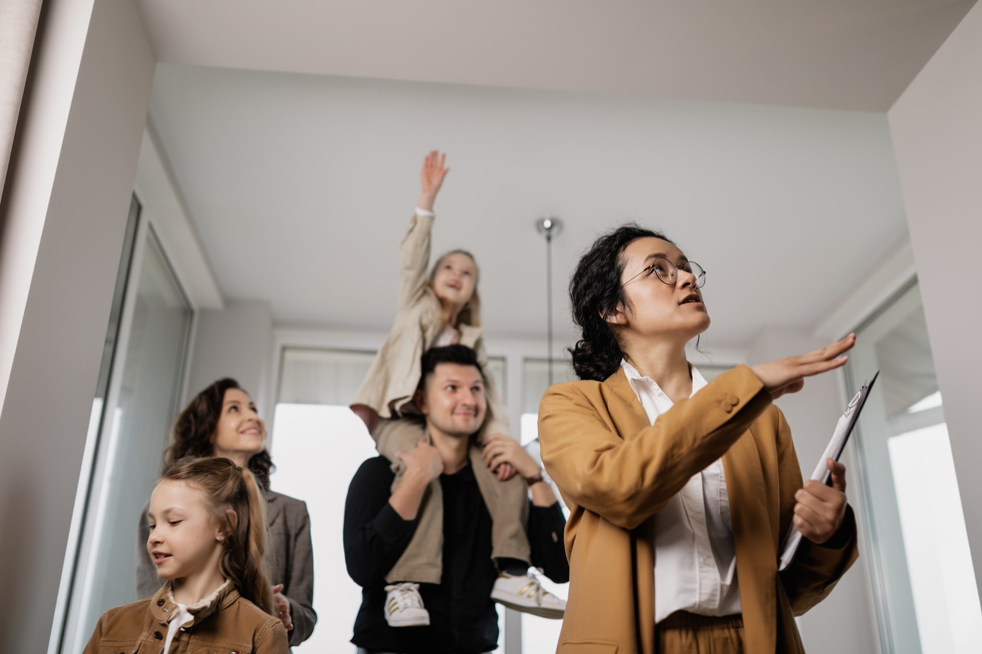 A realtor in a mustard yellow cardigan holding a clipboard shows a family of four around a home. The realtor is gesturing to the walls. The family is a mother, father, and two young daughters. The youngest is on the father's shoulders and reaches up playfully toward the ceiling as the mother watches and smiles. The other daughter inspects the opposite wall.