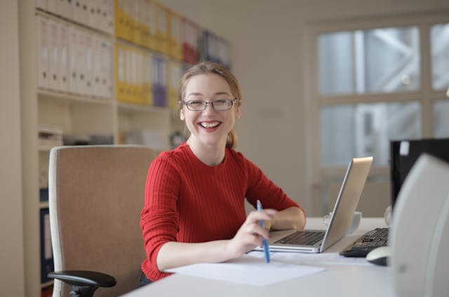 A woman in a red sweater smiles at the camera. She wears glasses and her blonde hair is in a ponytail. The woman is sitting at a desk in an office. Behind her along the wall are rows of binders.