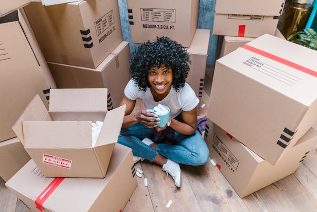 A young woman with short, curly black hair sits cross-legged on a wooden floor. She is holding a light blue mug filled with packing peanuts, and is surrounded by stacks of moving boxes.