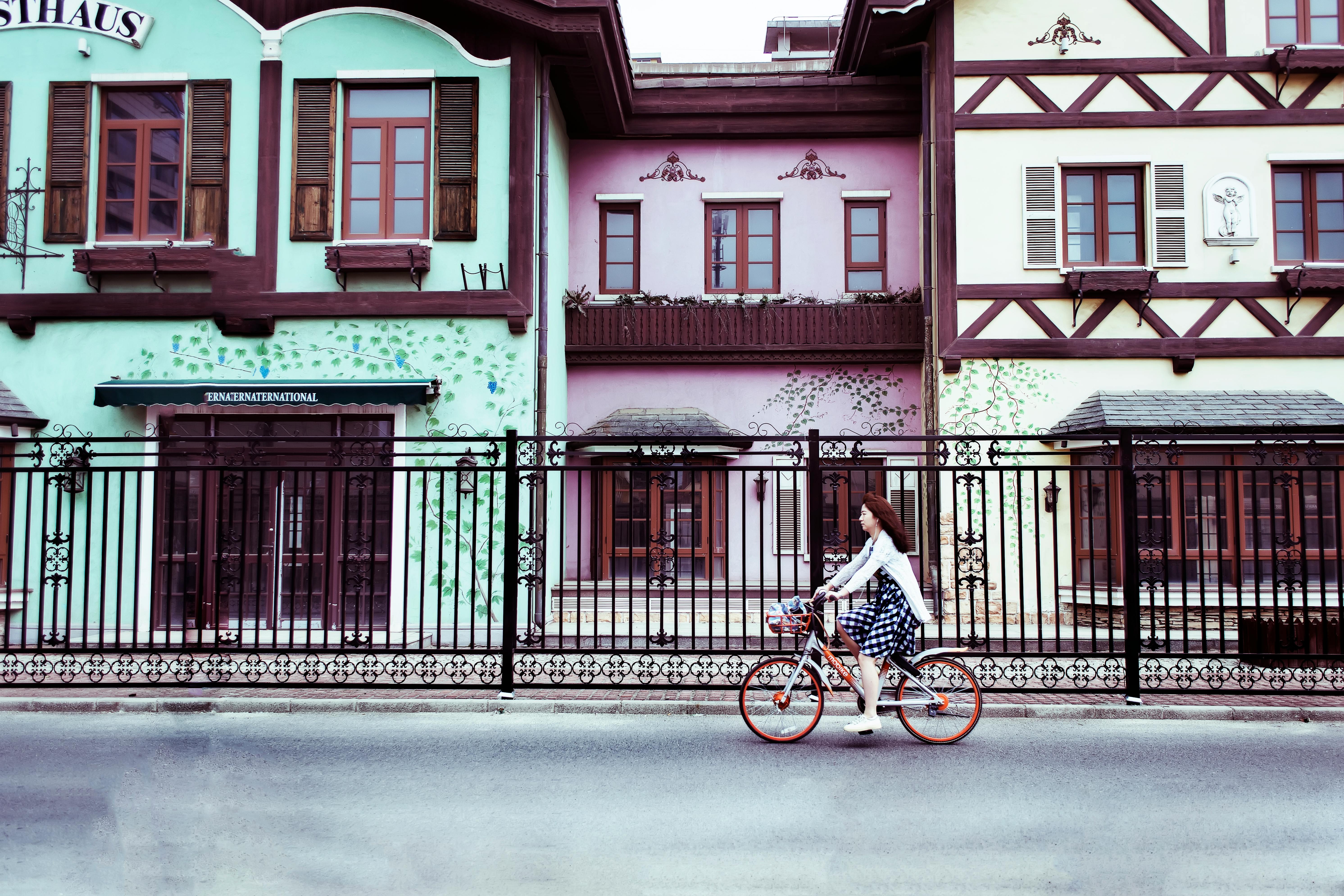 A woman rides a bike along a road. Along the road are three shops with apartments above them. A large metal fence separates the road and the shops.