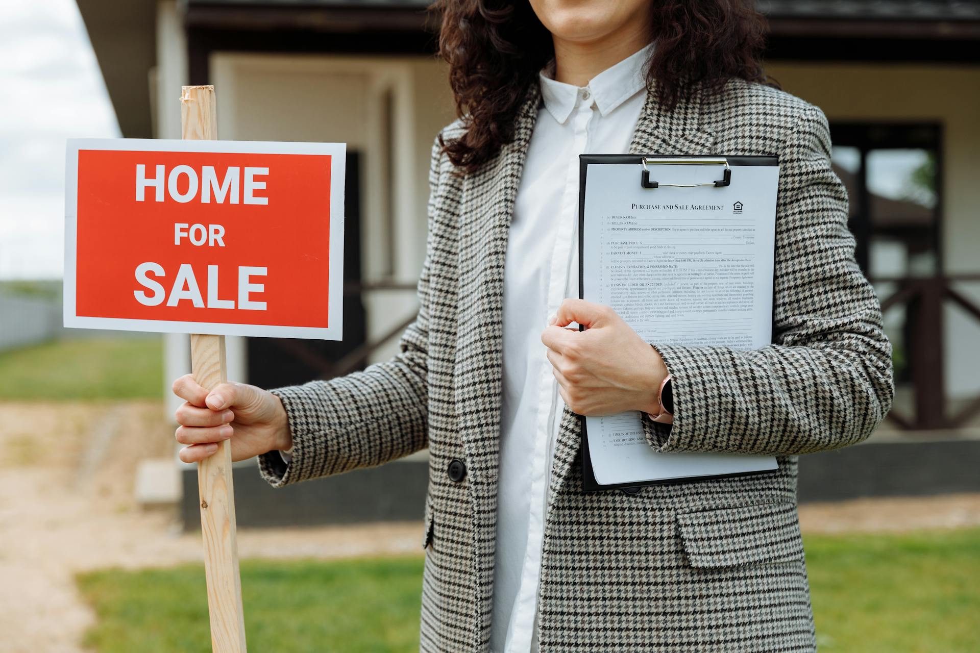 A woman in a plaid patterned grey blazer stands in front of a home. In one hand she holds a red sign that states home for sale. The other hand holds a clipboard with a real estate form to her chest.
