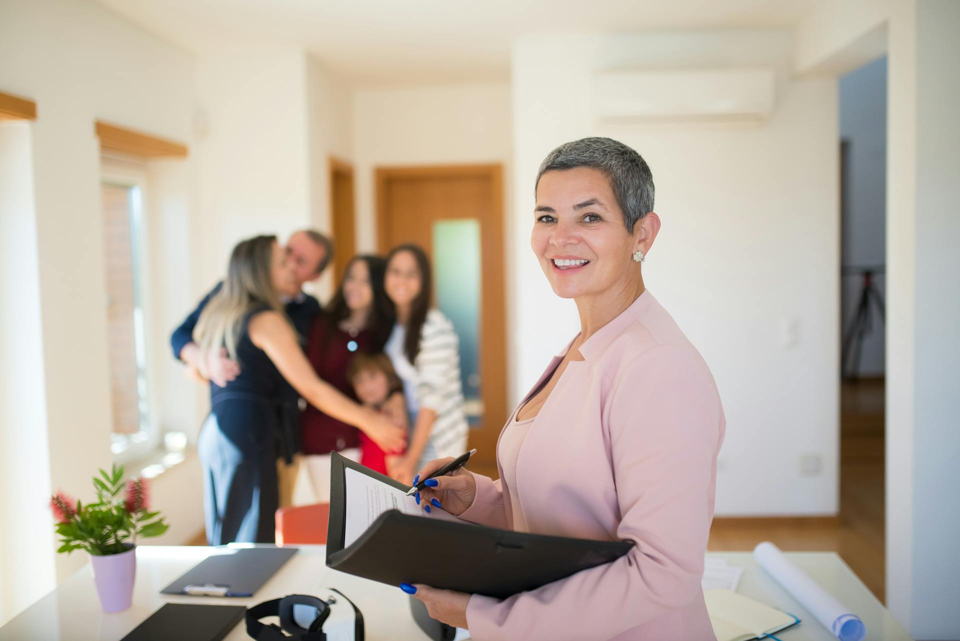 A woman with very short salt and pepper hair wearing a dusty pink blazer and holding a black portfolio stands in the foreground, smiling at the camera. In the background is a family of 5 happily embracing in a warmly lit room.
