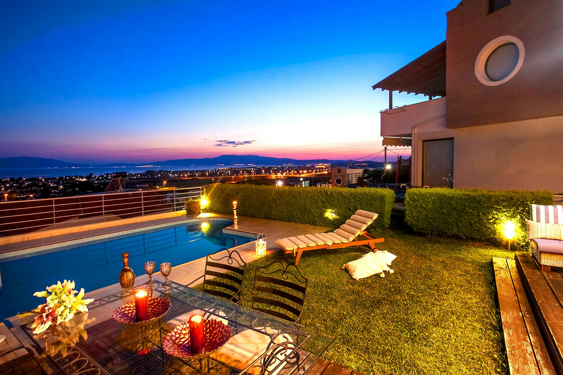A photo of a beautiful yard at sunset. The background is an expansive view of a distant city in a valley. The yard contains a small pool and deck chair, as well as a glass table with embellished metal chairs.