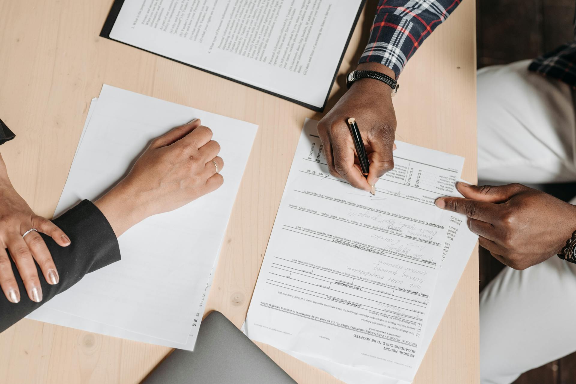 An overhead photo of a person's hands as they sign a form. Another pair of hands rests on the opposite side of the desk.