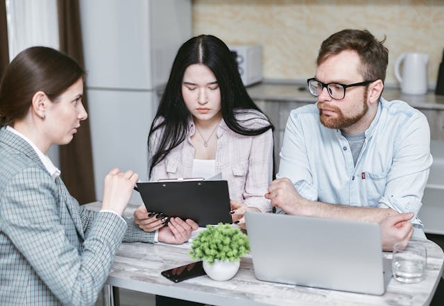 A man and a woman sit in a kitchen opposite another woman. The lone woman presents the pair with a document on a clipboard as the duo looks on with dejected expressions.
