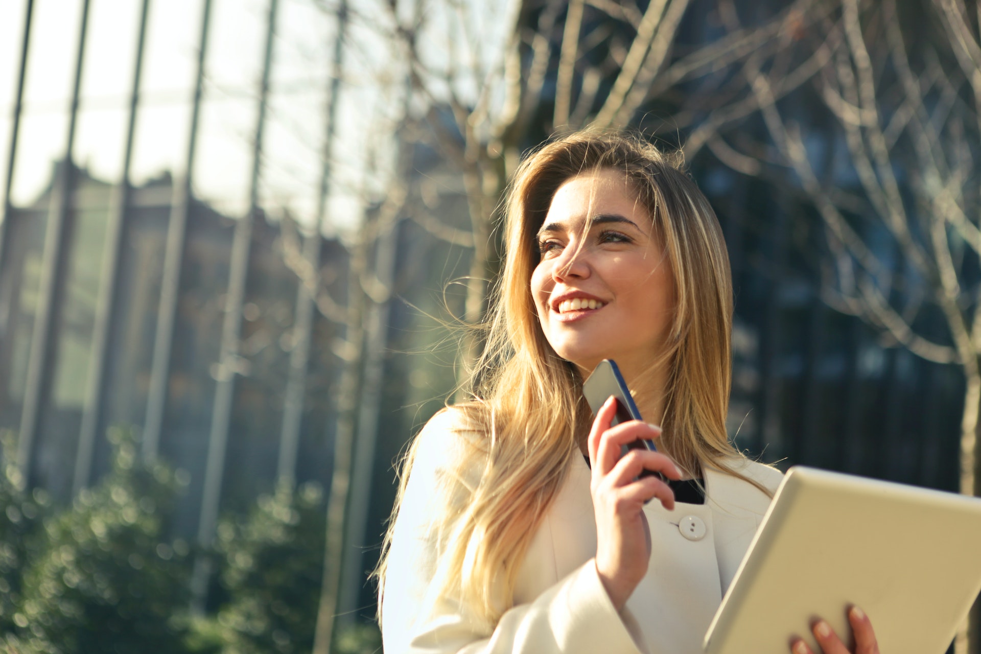 A woman in a cream jacket looks to the left of the photo and smiles. She is holding a phone in one hand and a tablet in the other. Behind her is an office building wall of windows, and various trees.