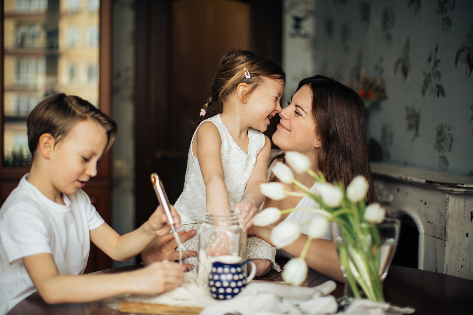 A family of three sit around a small kitchen table. The mother holds a young girl in her lap as the two giggle and smile at one another, faces close. A young boy holds a whisk and plays with baking supplies on the table. There are white tulips in a clear vase.